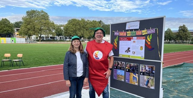 A man dressed in a chili costume and a woman stand by an info board on a sports field